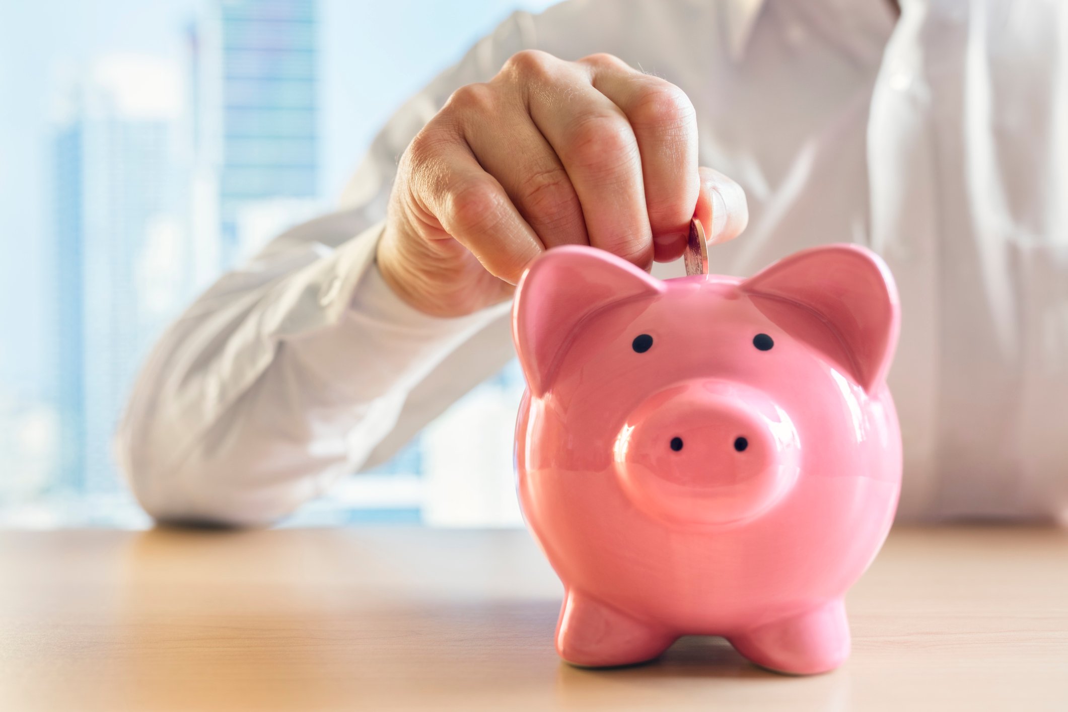 A person sitting places a coin into a piggy bank sitting on a table.