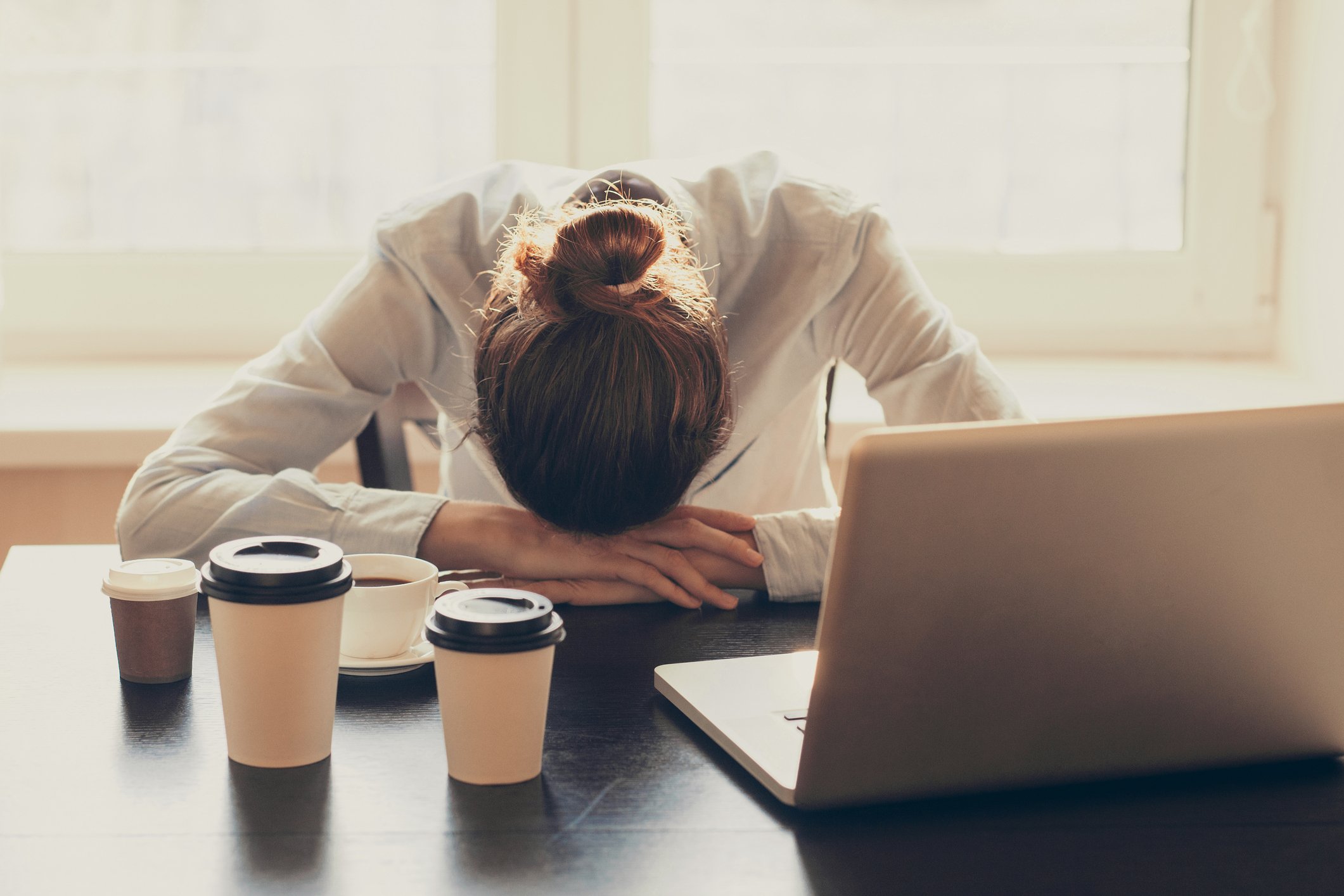 A woman has her head down on a desk with multiple cups of coffee and a laptop in front of her.
