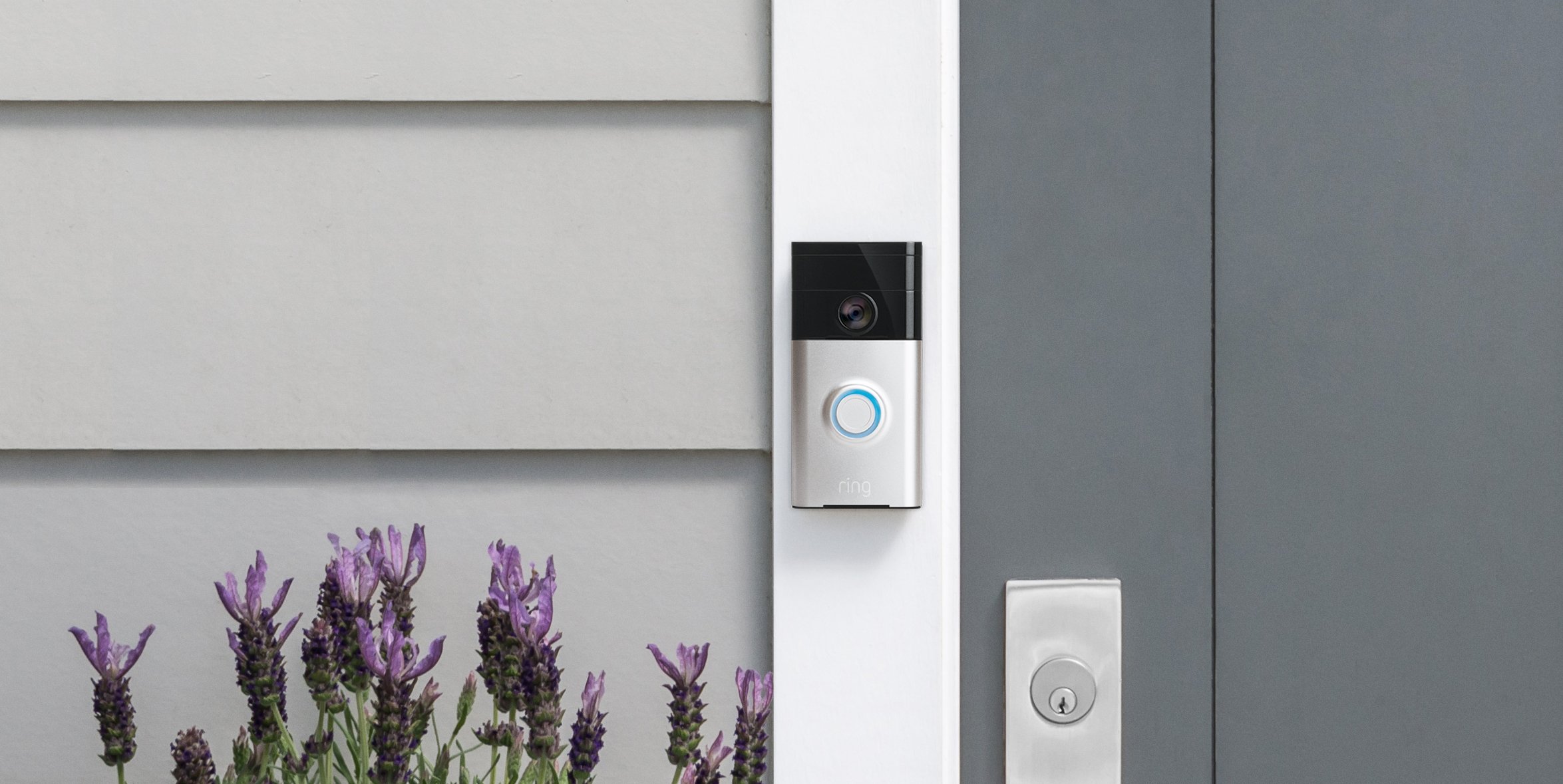 Ring Video Doorbell pictures attacked to the door frame of the front door of a grey house with purple flowers below it