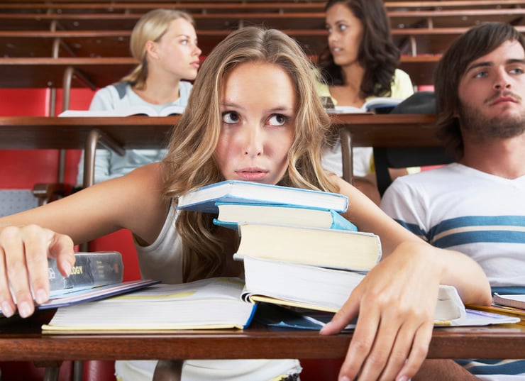 College student in lecture hall with chin on stack of textbooks, looking overwhelmed.