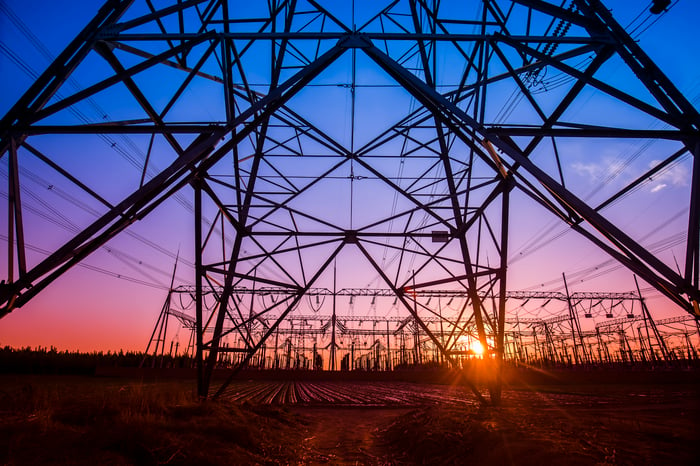 An electricity transmission pylon under twilight skies of blue, purple, and orange with the sun setting in the distance