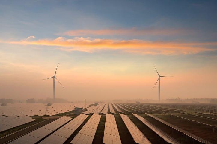 Rows of solar panels with two wind turbines in the distance, under a blue sky.