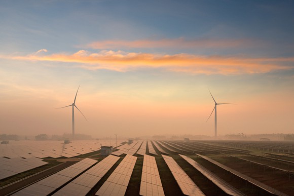 Rows of solar panels with two wind turbines in the distance, under a blue sky.