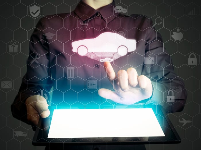 A man holding a tablet computer. Above the tablet, a simple image of a car floats in the air amid various icons related to money, shopping, and communications. He is clicking on the car image..