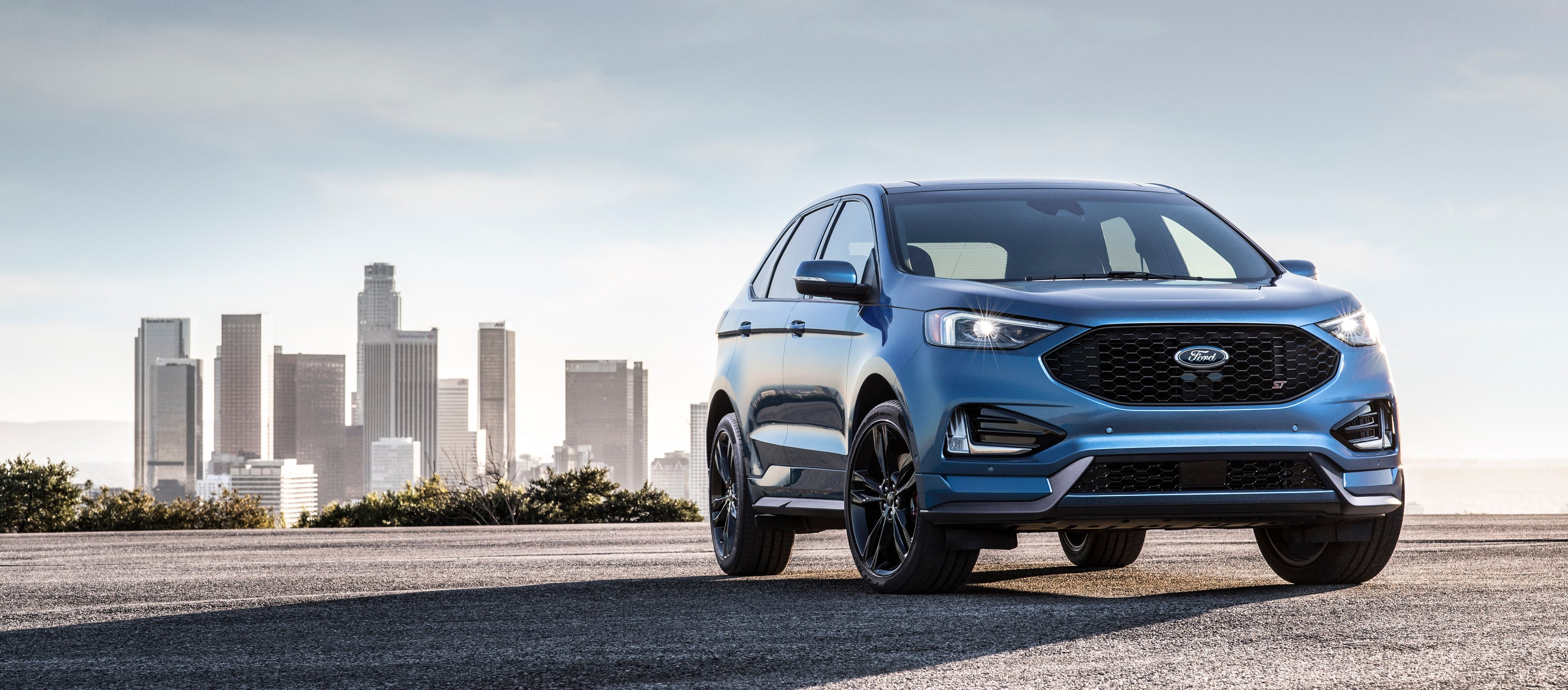 A blue 2019 Ford Edge ST, a midsize high performance SUV, is shown parked with a city skyline in the background. 