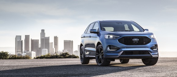 A blue 2019 Ford Edge ST, a midsize high performance SUV, is shown parked with a city skyline in the background. 