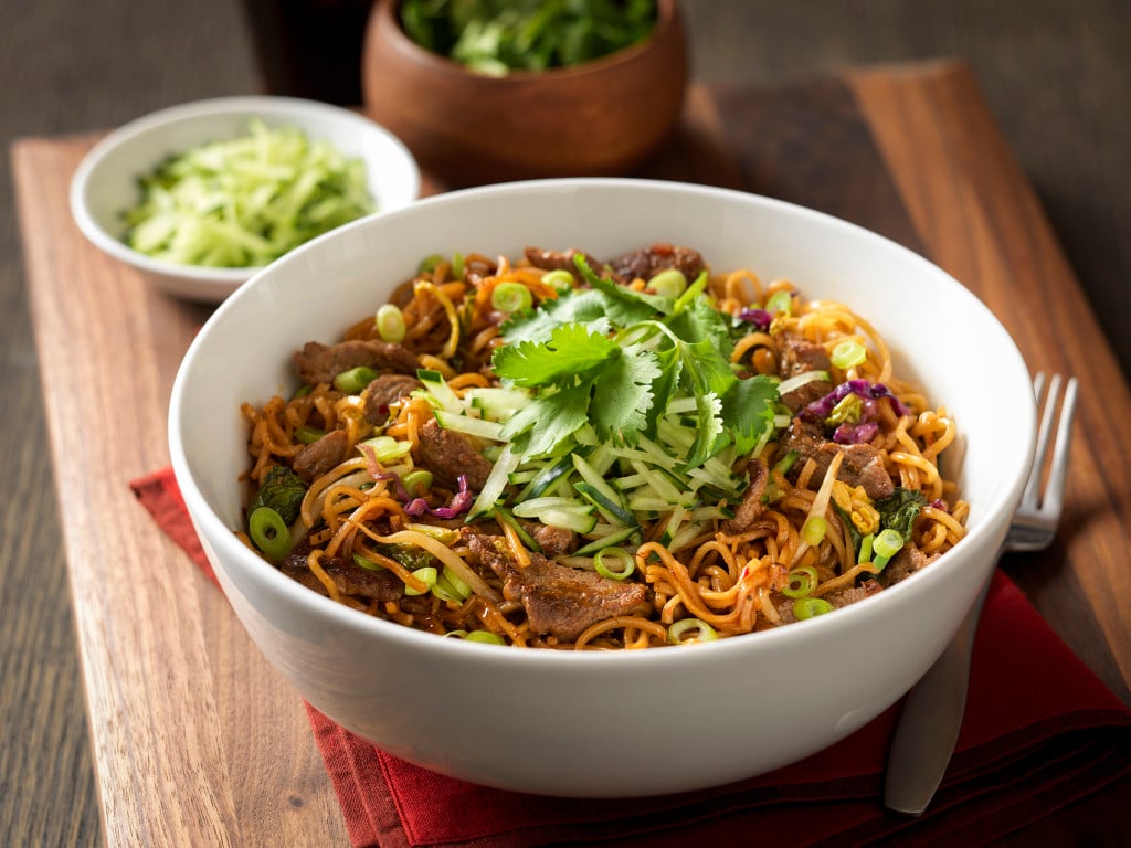 White bowl of noodles and beef with scallions and other ingredients, on a wood table with a bowl of salad and another side dish.
