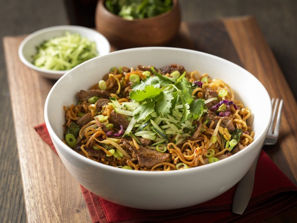 White bowl of noodles and beef with scallions and other ingredients, on a wood table with a bowl of salad and another side dish.