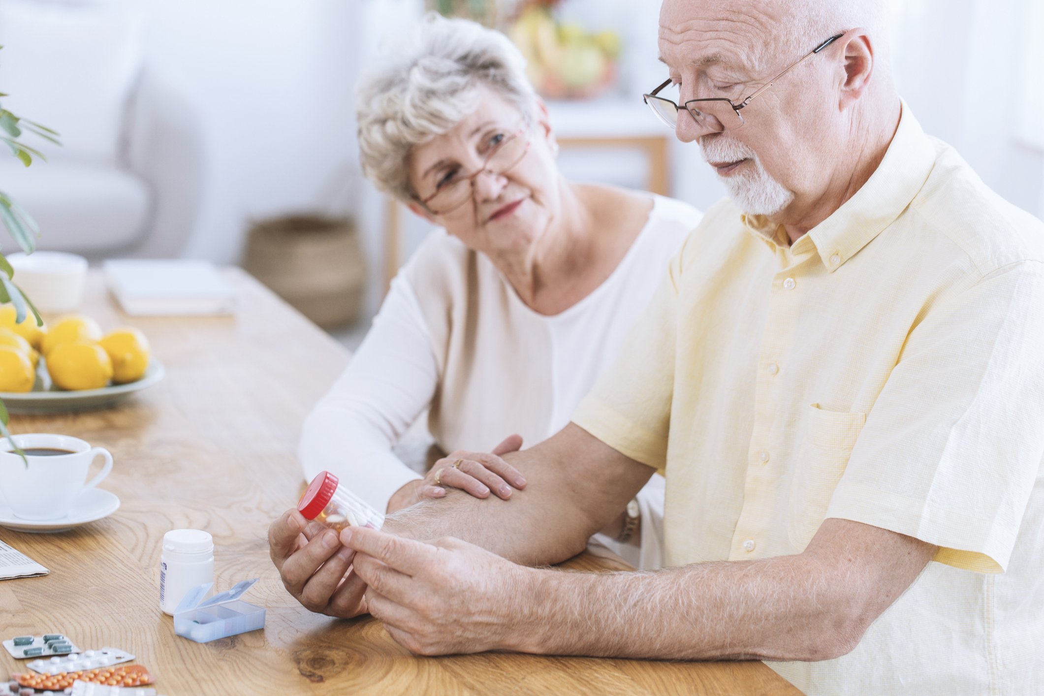 Elderly man and woman sitting at a table. The man is holding a pill bottle, and the woman is looking at him with her hand on his forearm.