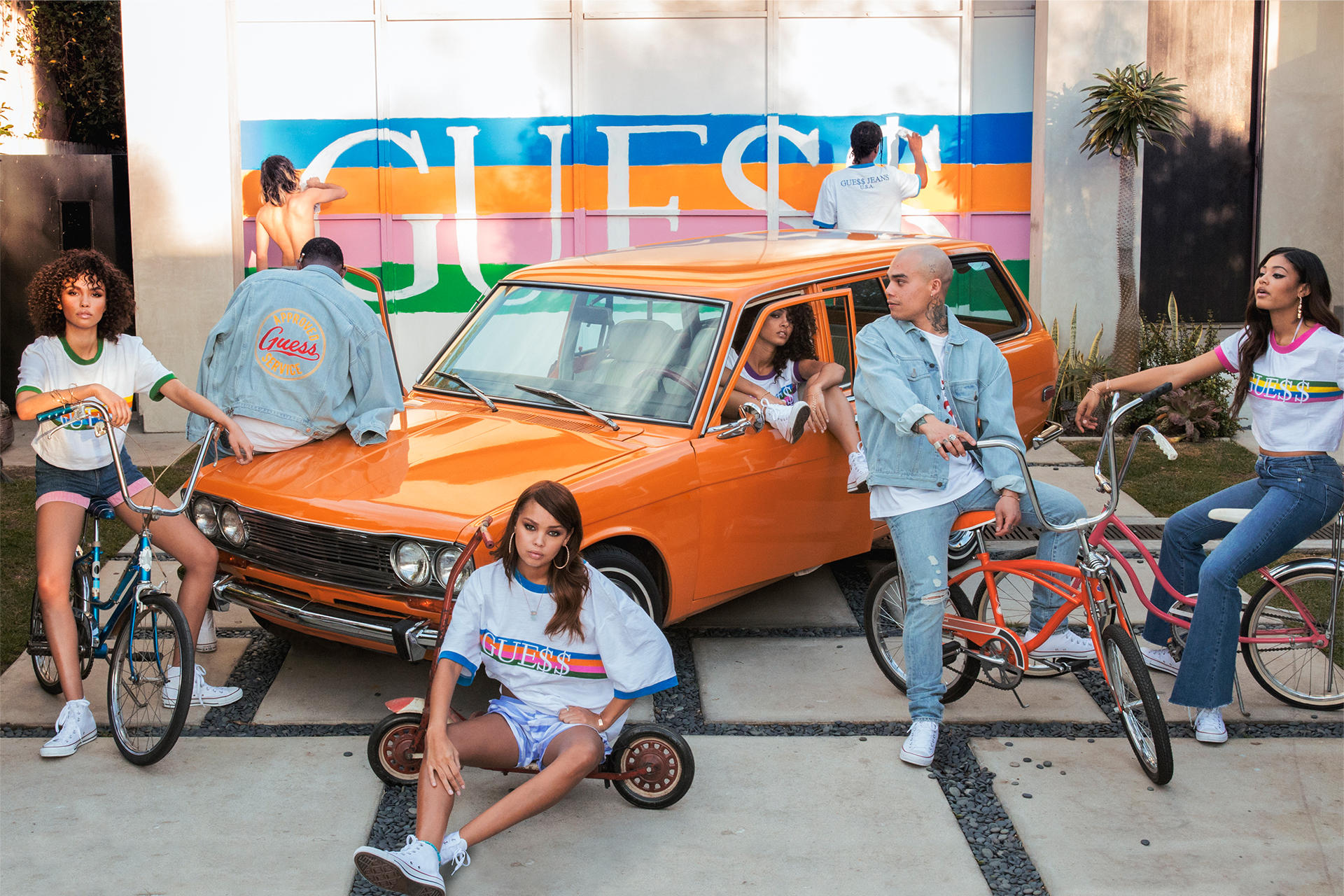Six fashion models sitting on bicycles in front of an orange car with the Guess logo painted on a building wall.