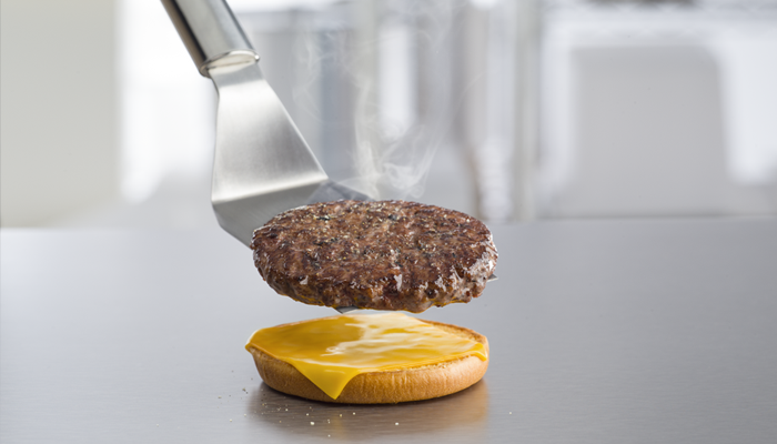 Sizzling beef patty on spatula hovering above the first layer of a Quarter Pounder with cheese.