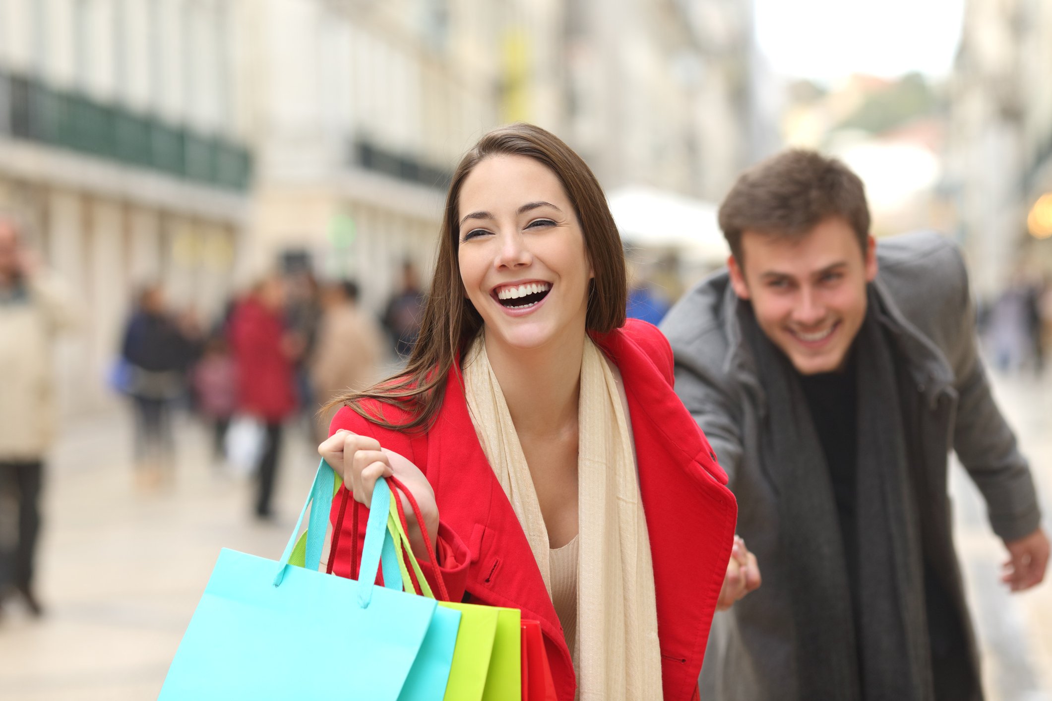 Two smiling young people carry shopping bags in a crowded city street.