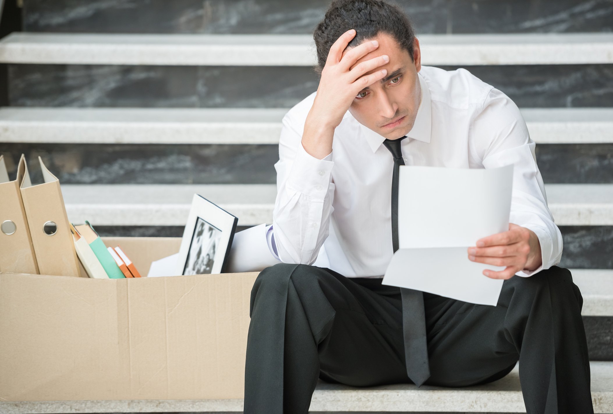 Man clutching his head while holding a piece of paper and sitting next to a box of desk supplies