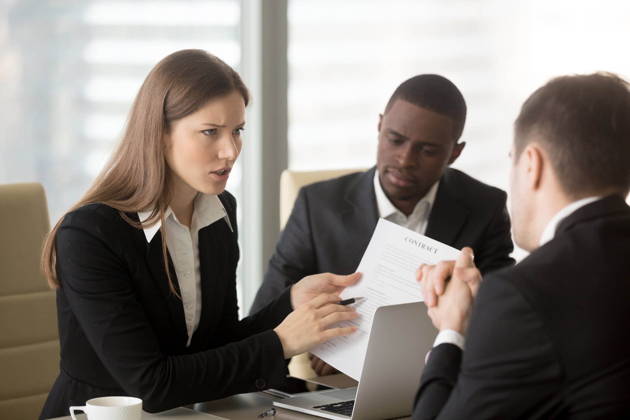 In an office setting, people in business attire review a contract, with a woman pointing to the document with a look of concern.