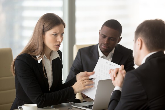 In an office setting, people in business attire review a contract, with a woman pointing to the document with a look of concern.