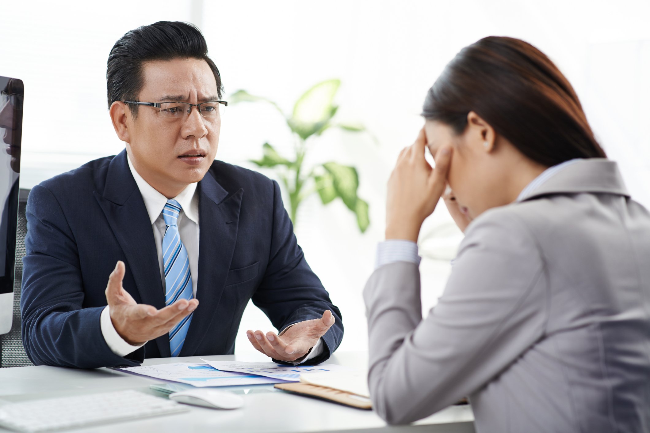 Man in suit gesturing toward woman in suit sitting across from him and holding her head as if crying