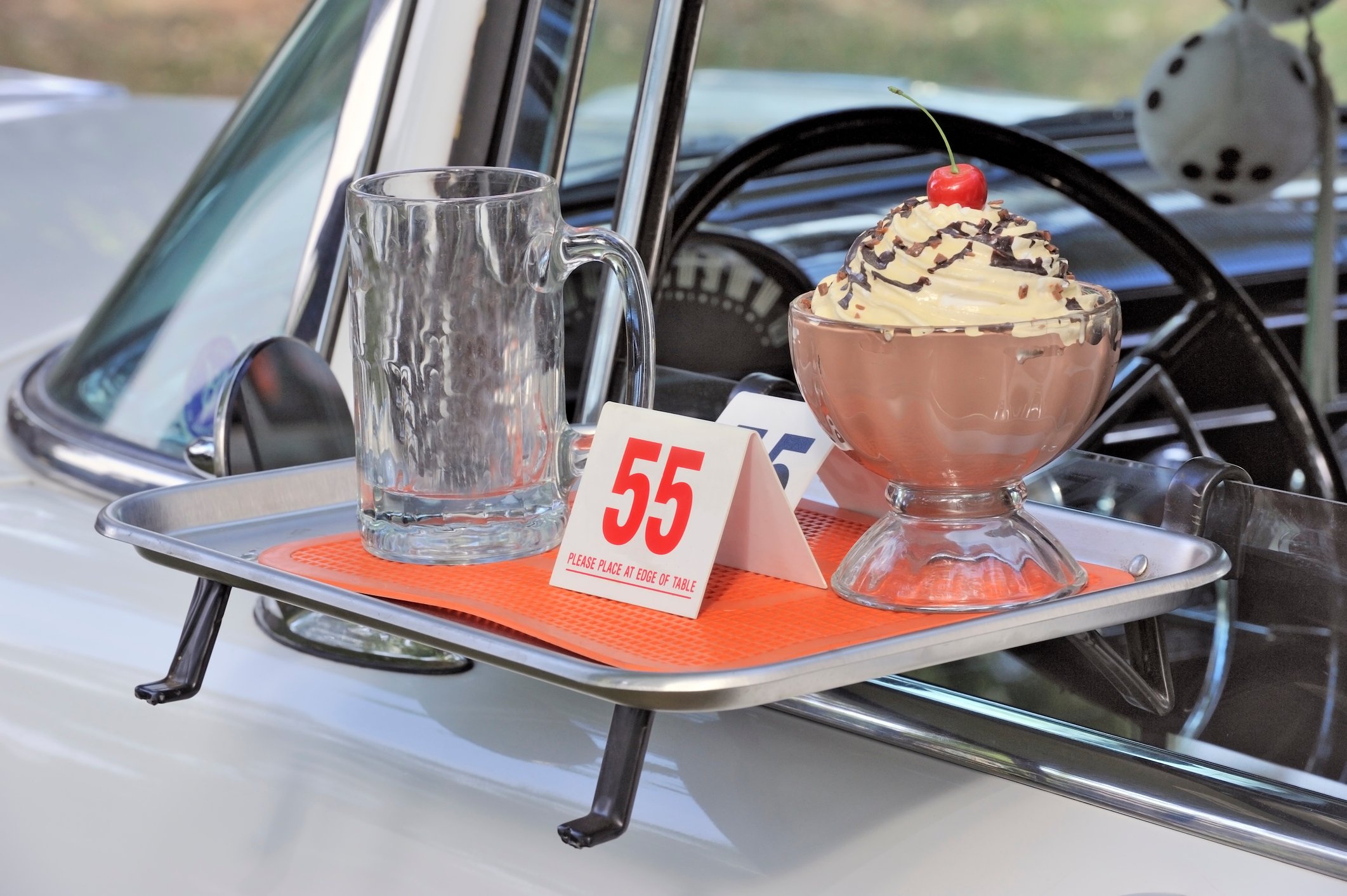 A tray golding an empty glass and a bowl of pudding on a car window at a drive-in.