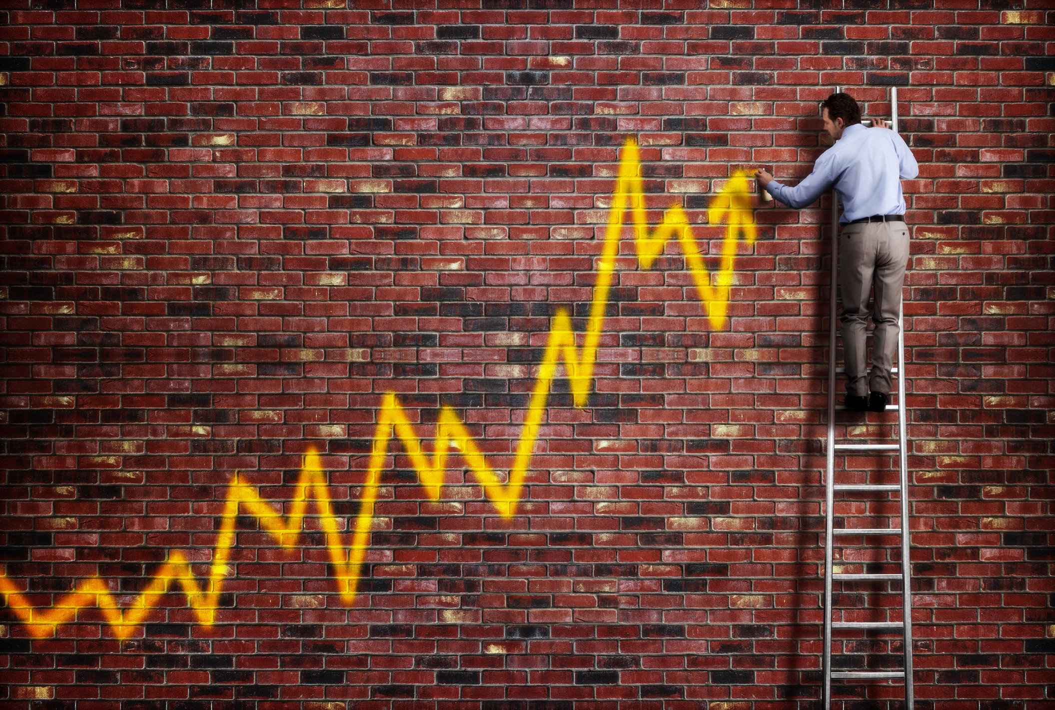 Man in business attire on a ladder drawing a chart on a brick wall indicating gains.