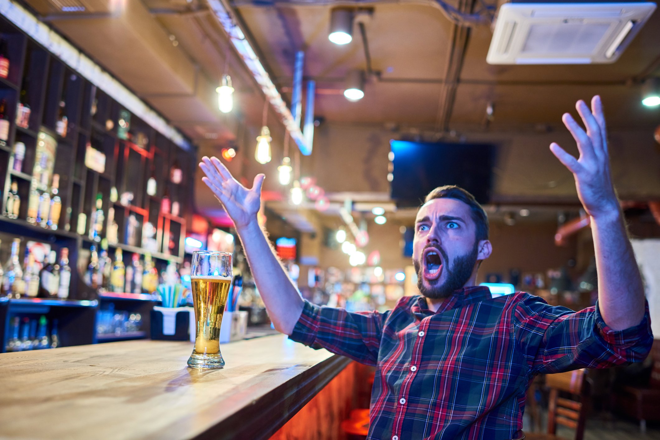 A bearded man sitting in a bar with his hands raised and his mouth open in, perhaps, shock.