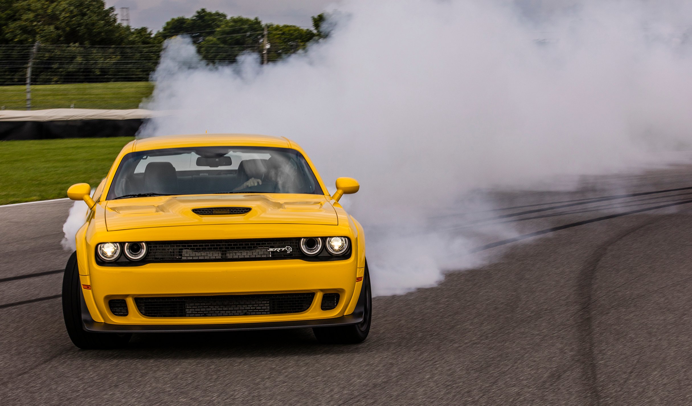 A yellow 2018 Dodge Challenger SRT Hellcat Widebody is shown powersliding around a racetrack with a cloud of tire smoke behind.