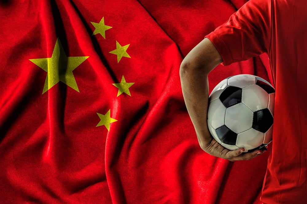 A man holds a soccer ball in front of a Chinese flag.