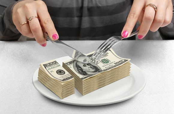 A woman uses knife and fork to cut up a pile of hundred-dollar bills on her dinner plate.