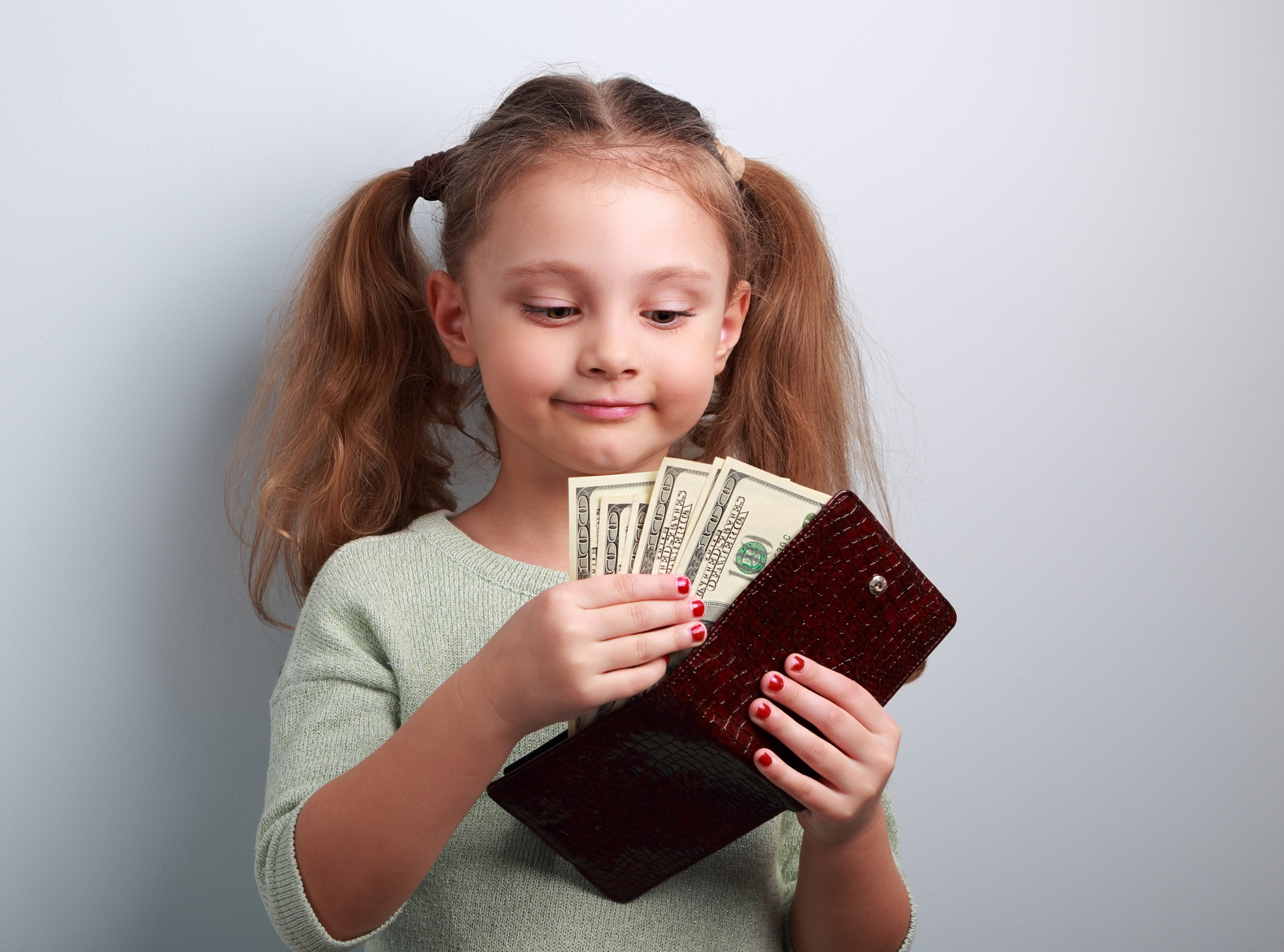A smiling little girl with pigtails and red nail polish counting dollar bills from inside a burgundy wallet 