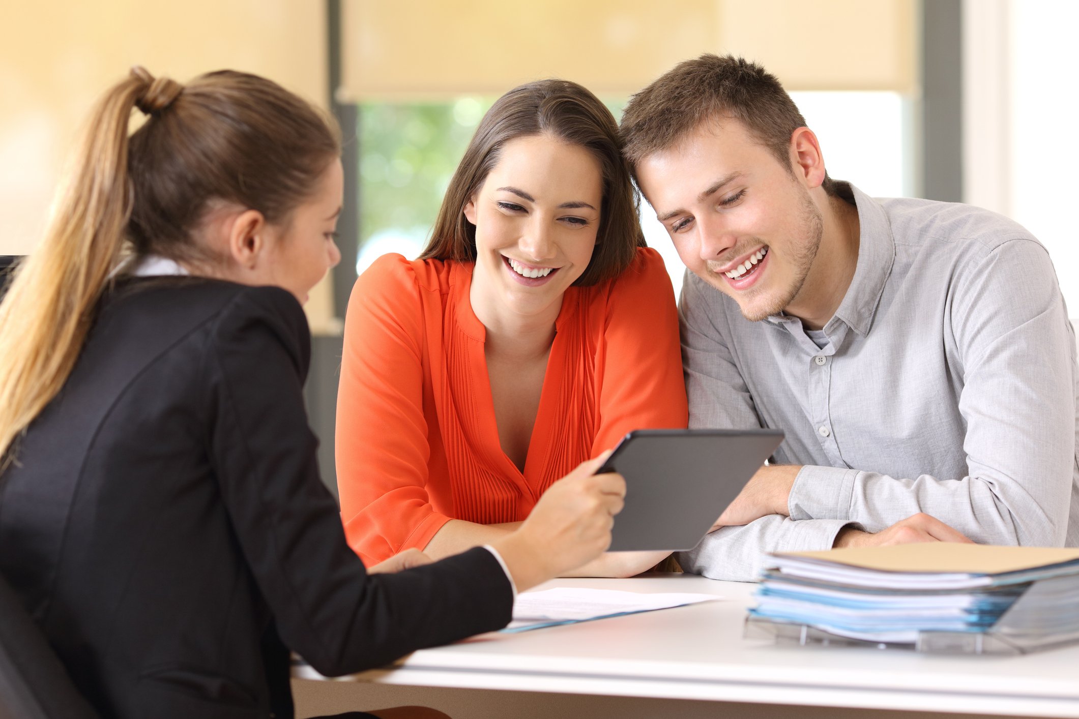 A smiling young couple closing the deal on a new home, with a female realtor and a large folder of paperwork on the table in front of them.