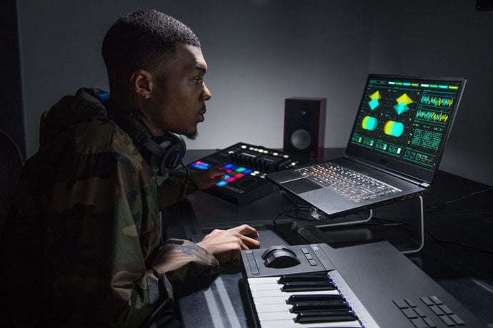 A man looking at a notebook computer elevated off a table with a stand, with a piano keyboard, sound mixer, and speaker on the table