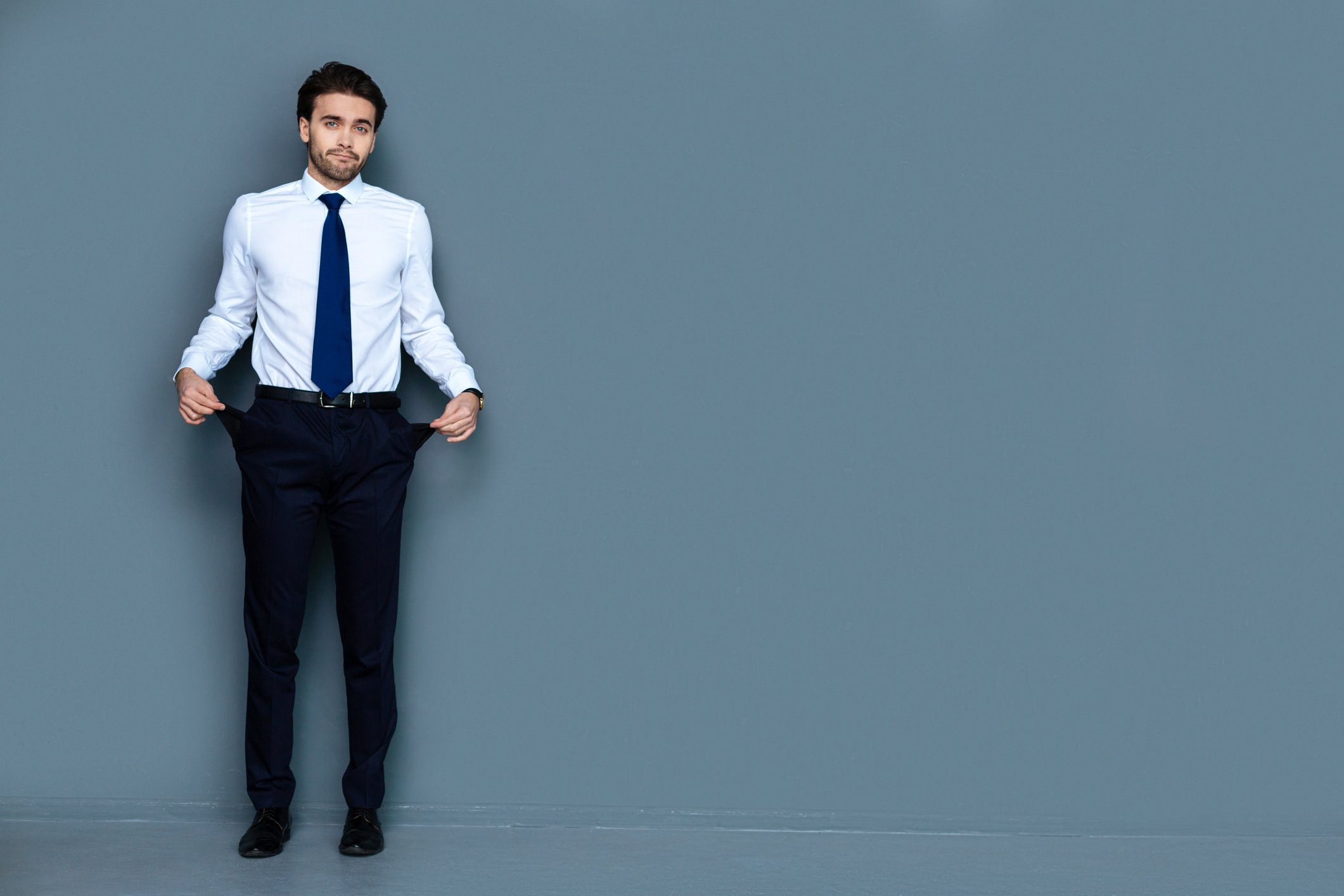 Man in white shirt, blue tie, black pants holding his pockets out to show that they are empty.