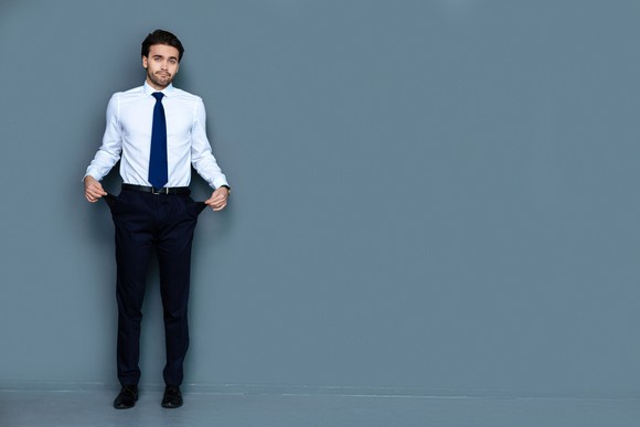 Man in white shirt, blue tie, black pants holding his pockets out to show that they are empty.
