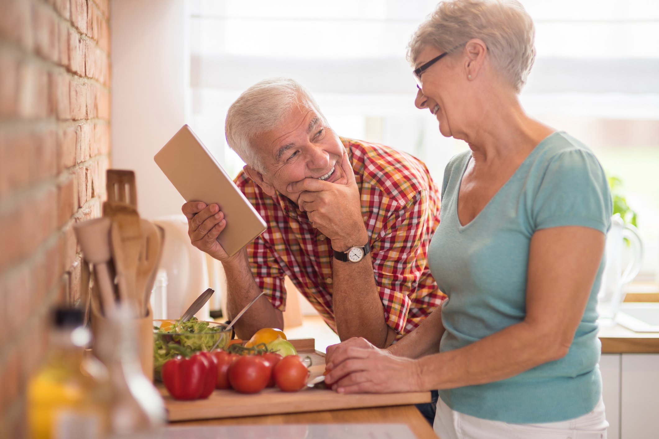 Senior man holding a notepad and smiling up at a senior woman while standing in a kitchen as she's cutting vegetables