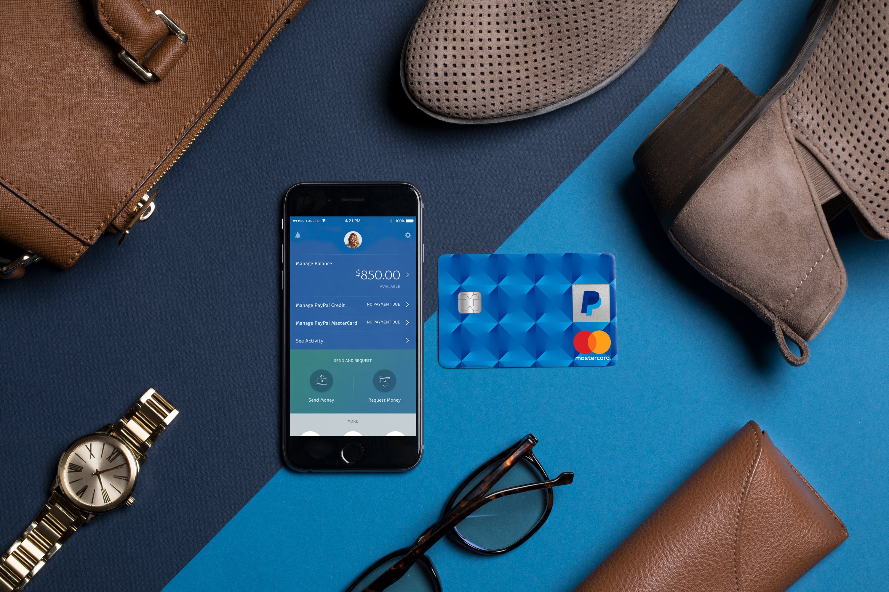 A phone displaying PayPal mobile app laying on a desk with a credit card, glasses, shoes, watch, and other luxury items.