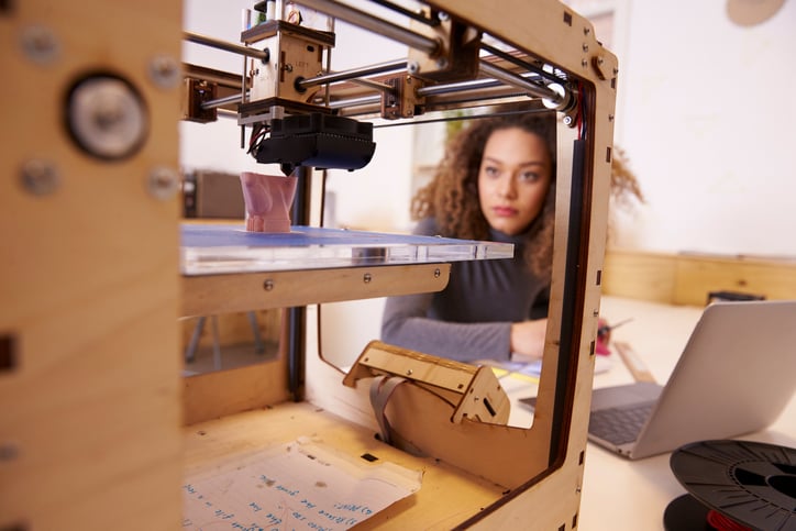 A woman on a laptop looking at a design in a 3D printer.
