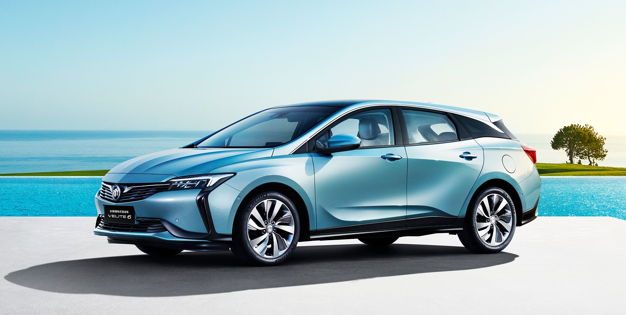 A blue Buick Velite 6, an all-new vehicle shaped somewhat like a station wagon. It's shown parked on a sun-splashed beachfront. 