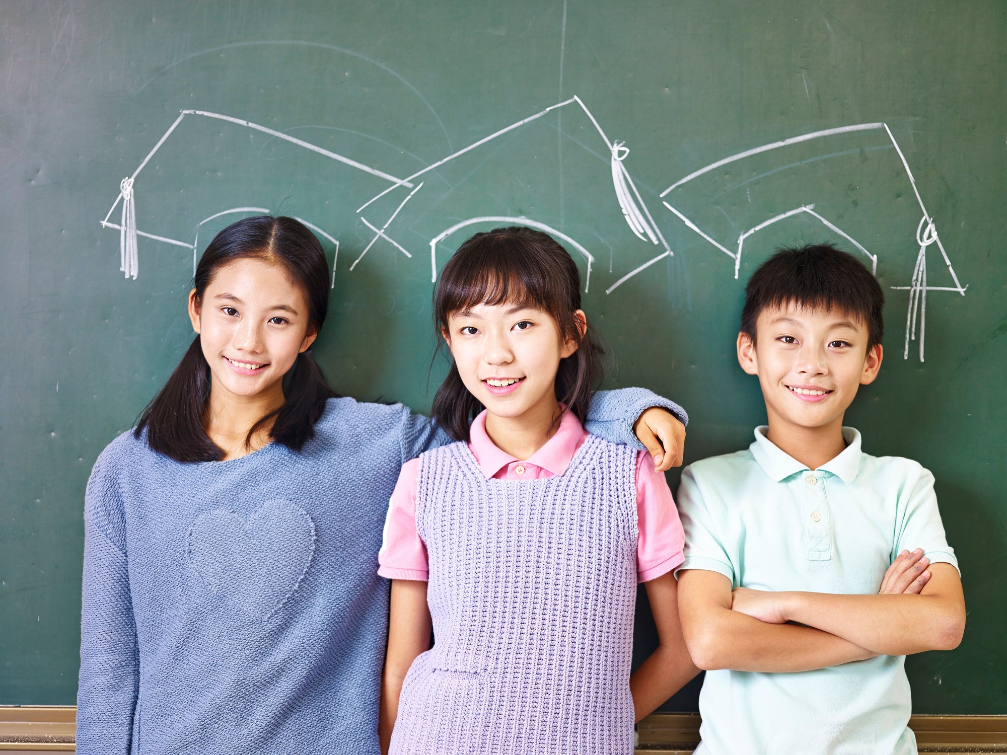 Three Asian elementary school children standing in front of chalkboard underneath chalk-drawn mortarboards