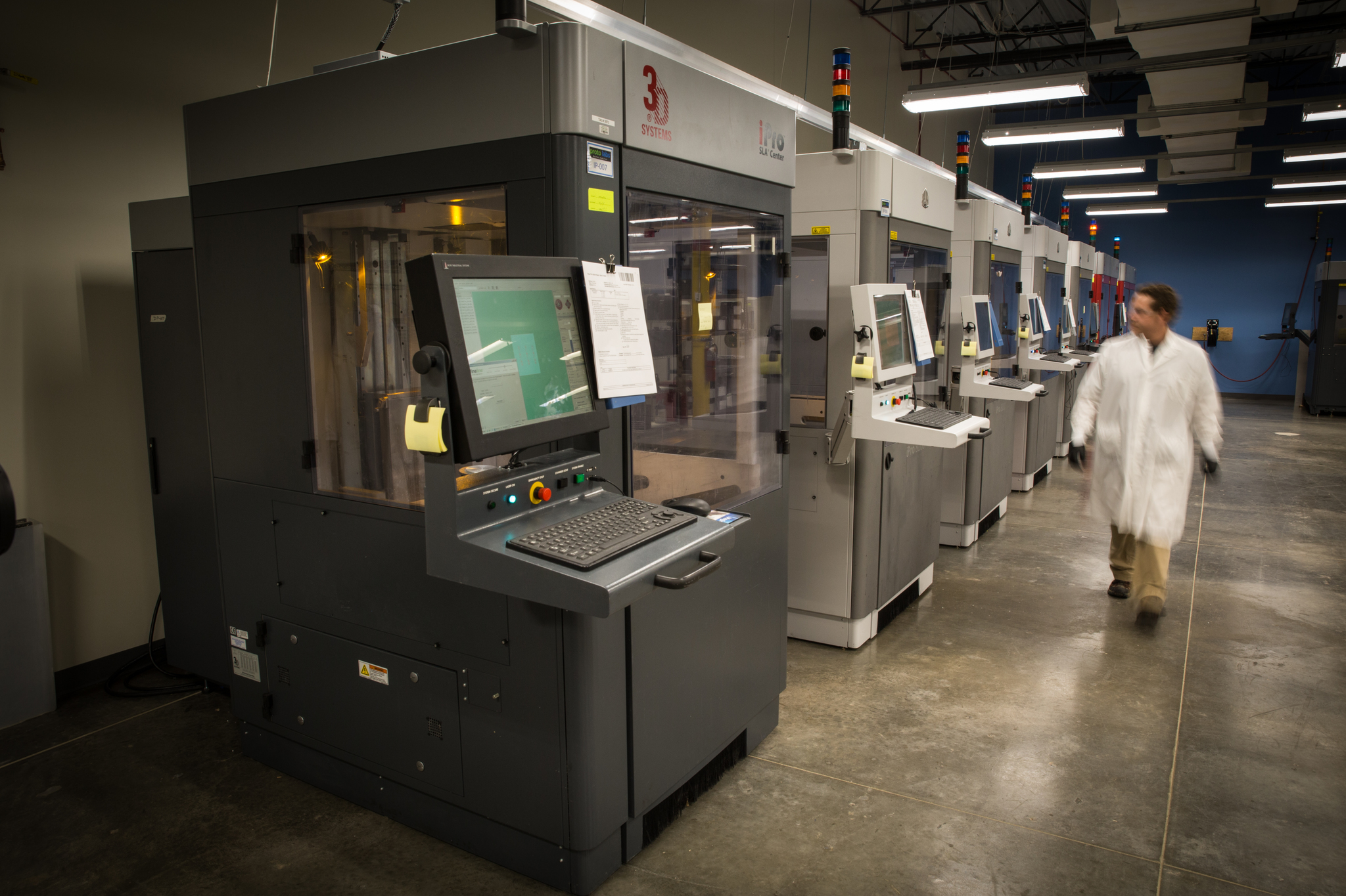 A worker walking by a row of industrial 3D printers in a Proto Labs' facility.