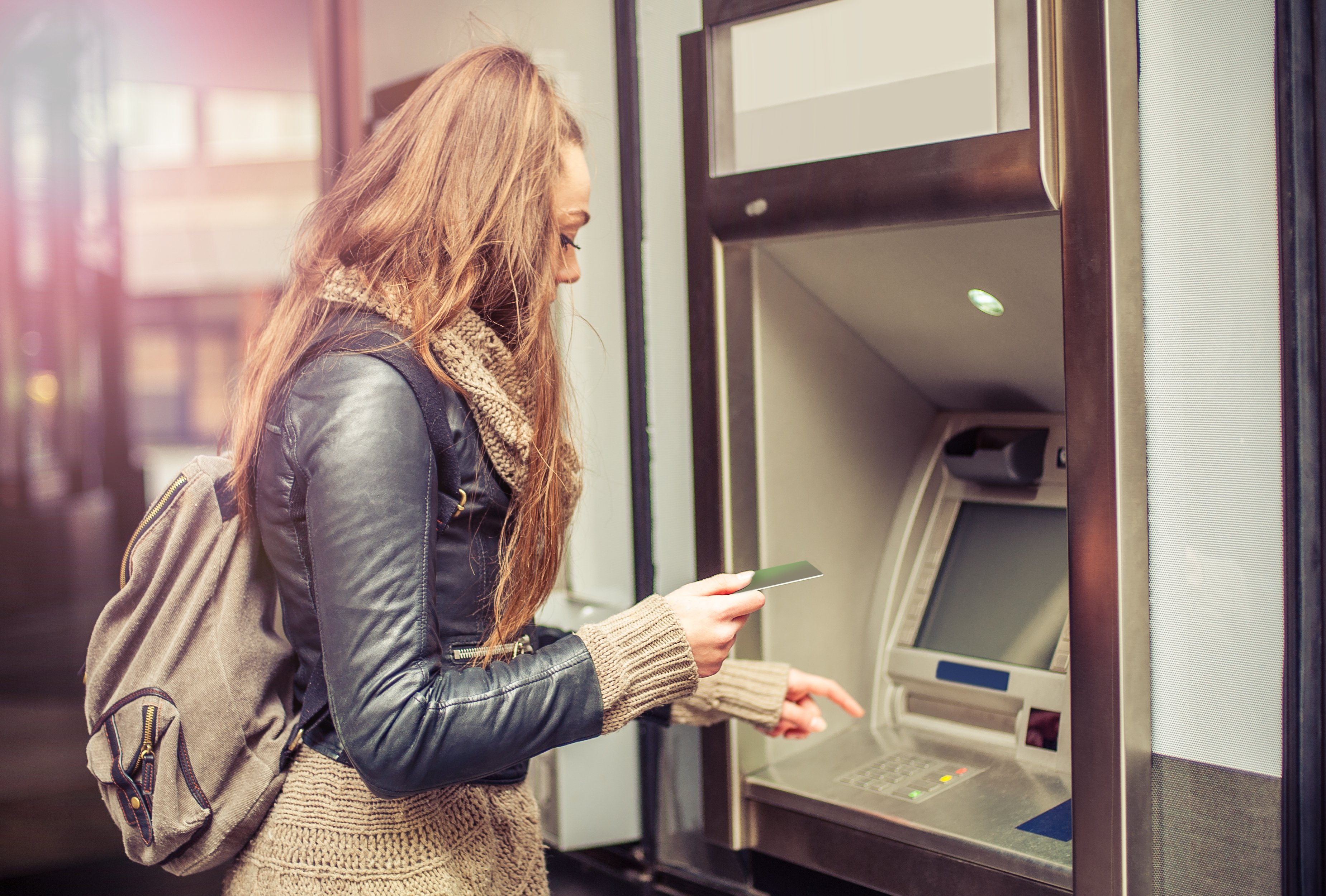 Young woman with backpack standing at ATM making withdrawal