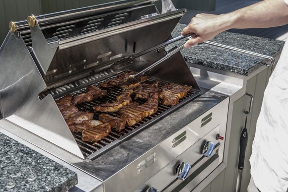 A man holds tongs over steaks on an outdoor propane grill