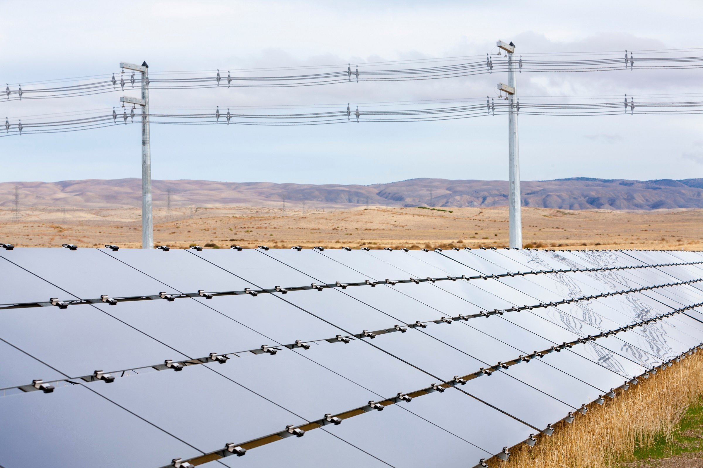 Four long rows of solar panels in a semi-arid landscape with power transmission lines in the background.
