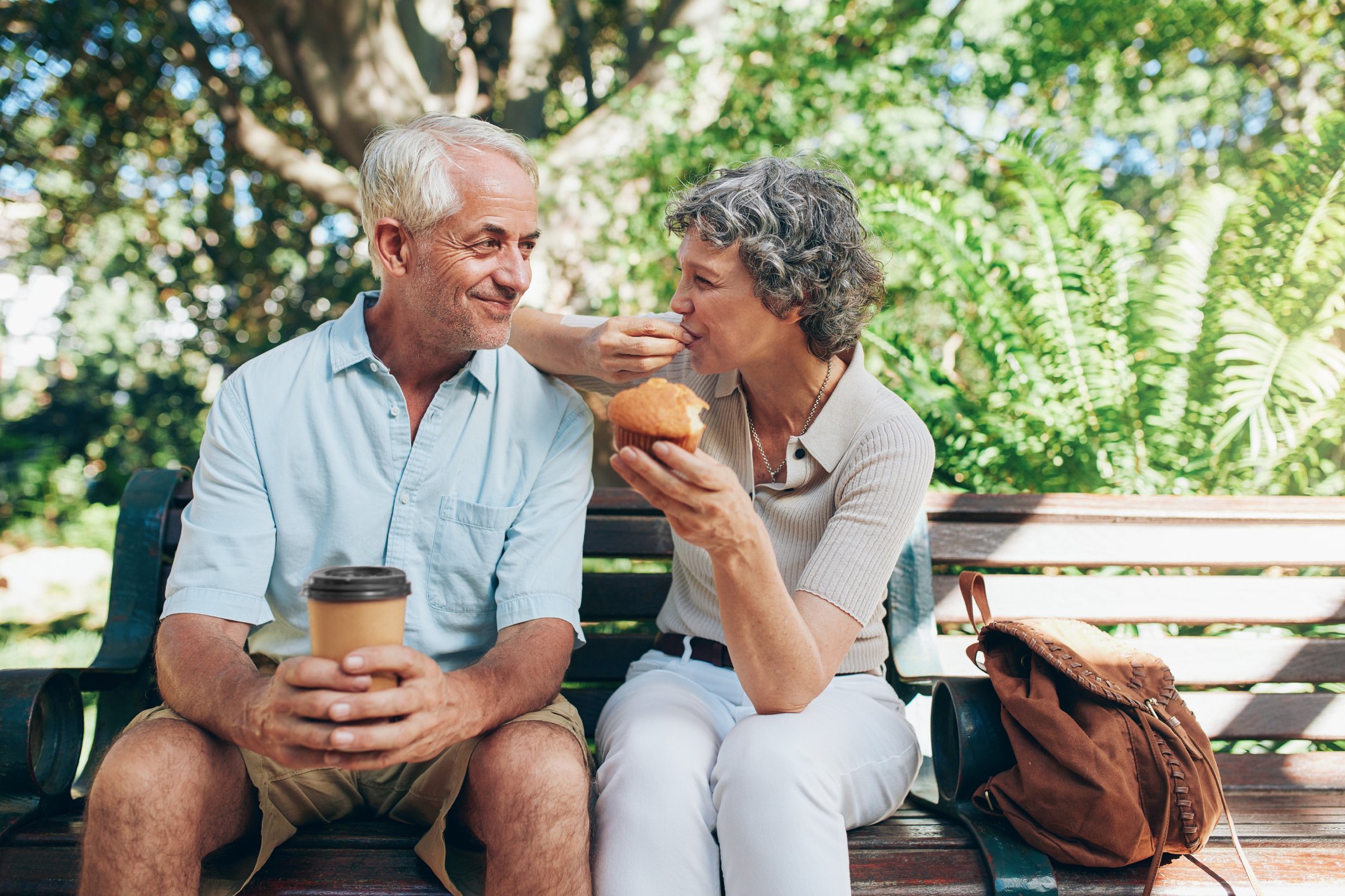 Senior couple on a park bench, with the male holding a cup of coffee and the female eating a muffin