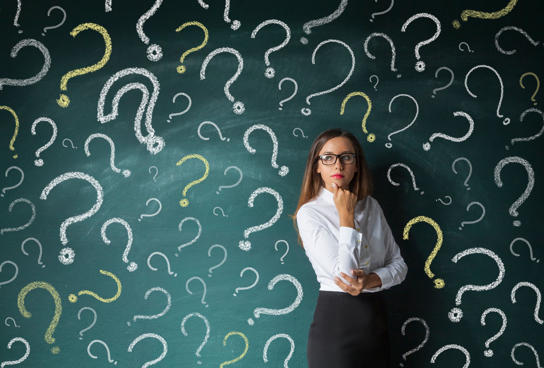 A businesswoman in a thinking pose while standing in front of a chalk board with many question marks drawn on it.