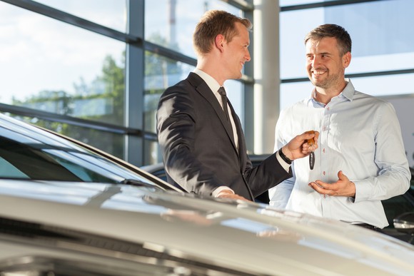 Person in suit handing key to smiling person next to a car inside a glass-walled building.
