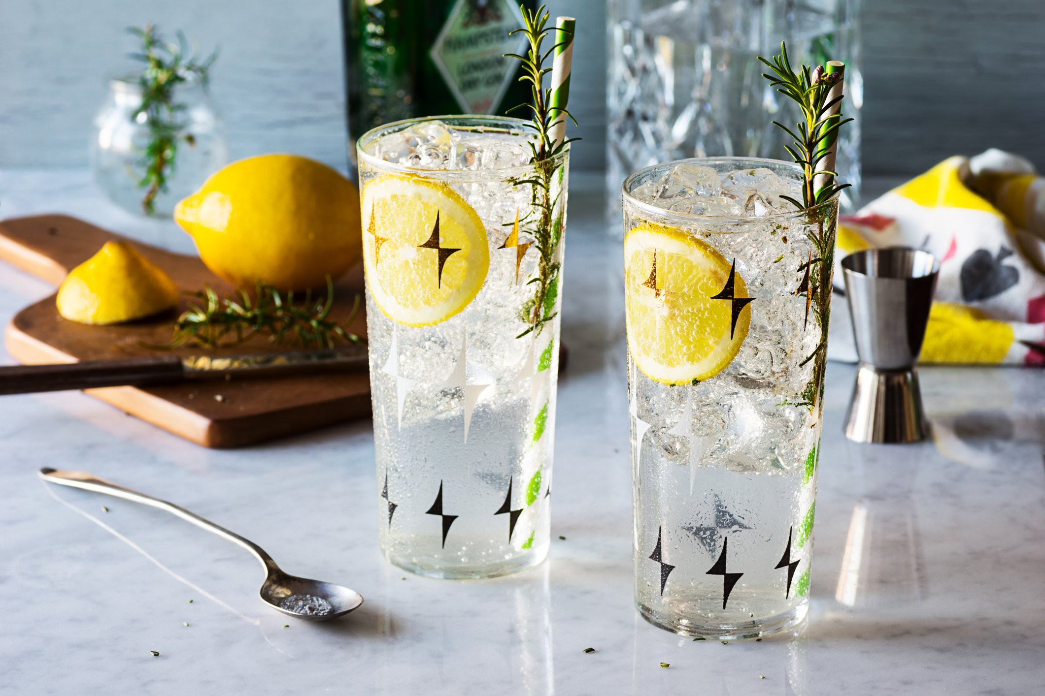 Two glasses with clear liquid and lemons sit on a counter with a lemon on a cutting board in the background.