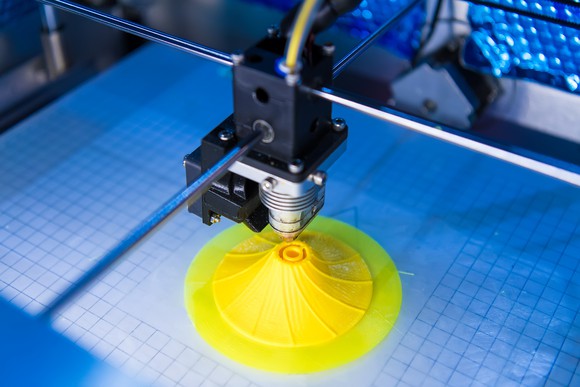 A 3D printer printing a yellow plastic object on a blue surface.