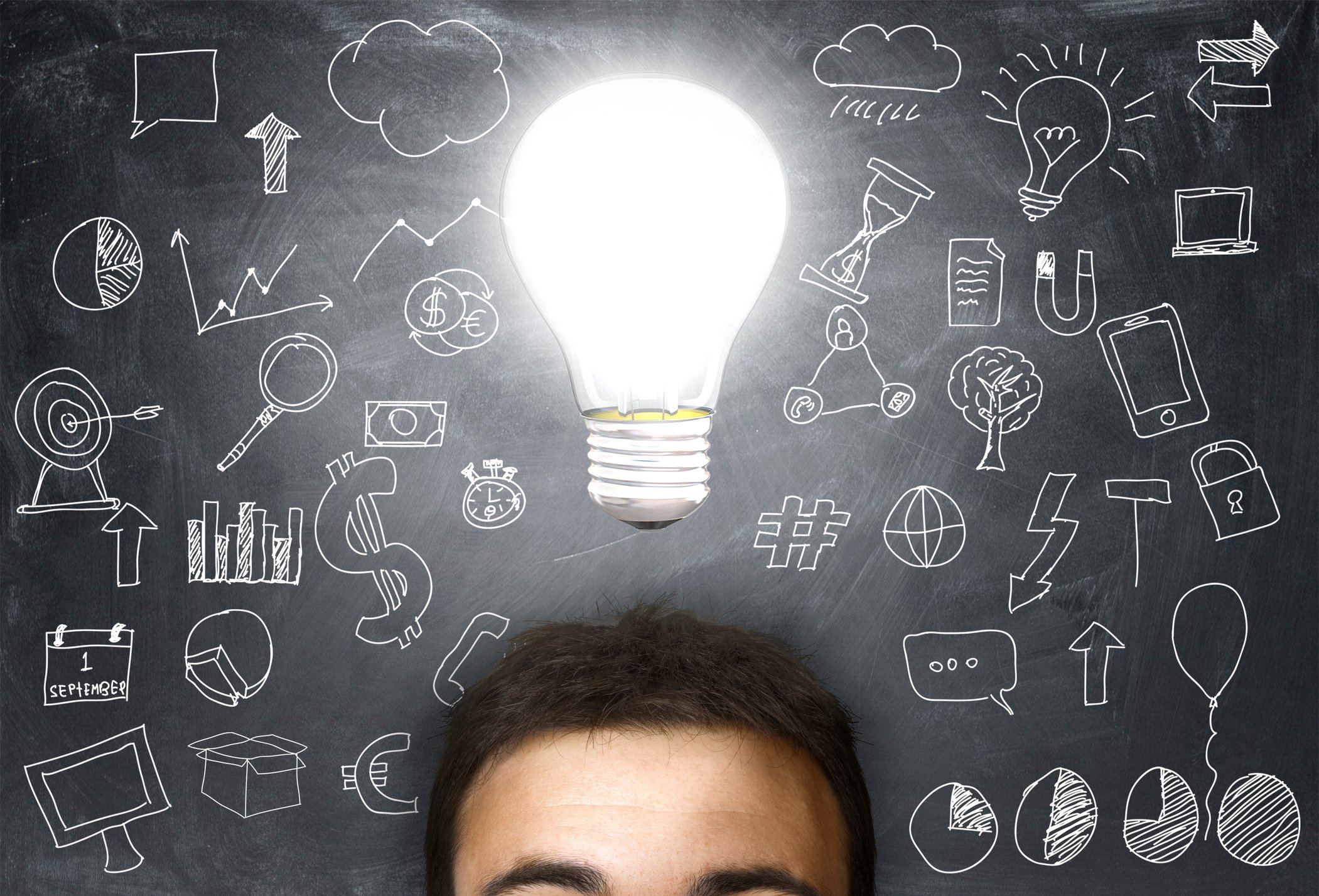 A view of a person's forehead with an illuminated lightbulb above it in front of a background showing a chalkboard covered by diagrams.