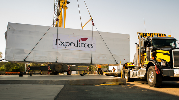 Crane carrying an Expeditors container with a semi trailer truck next to it.