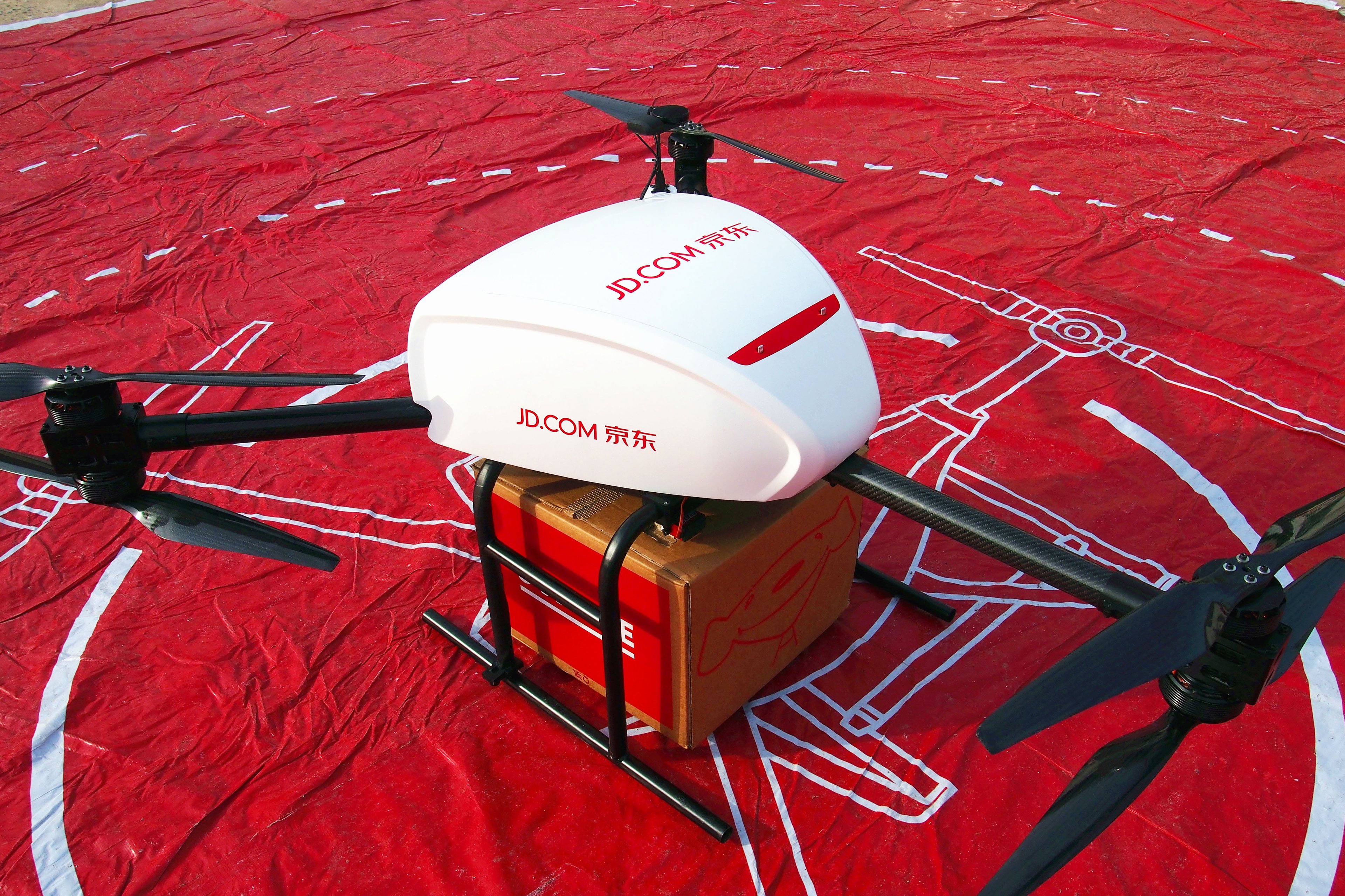 A delivery drone marked with JD's colors and logo, cradles a large cardboard box on a red tarp.