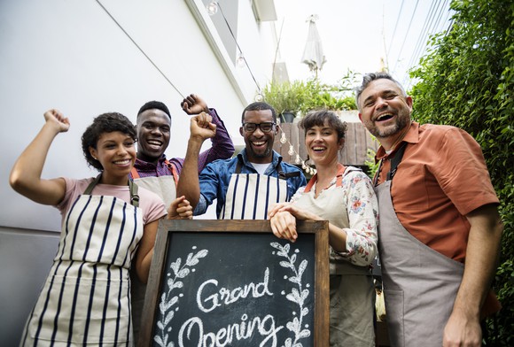 A group of people in aprons stand in front of a grand opening sign.
