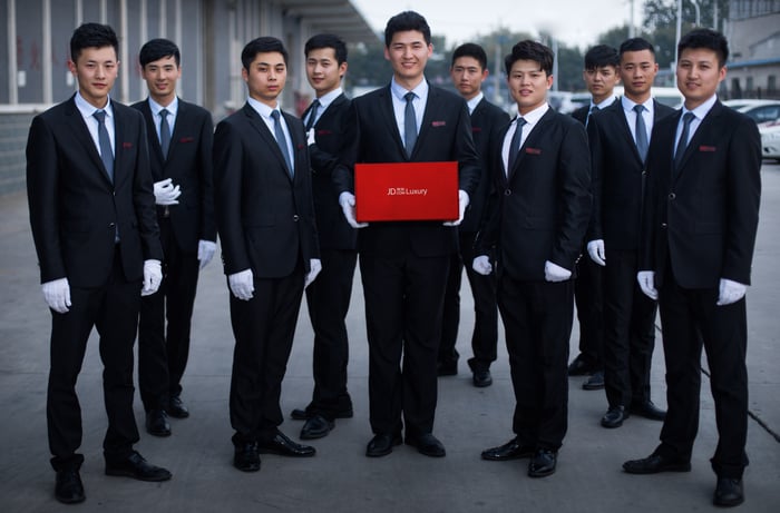 a group of young men in suits and white gloves face the camera as one holds  a red box labeled with JD's logo.